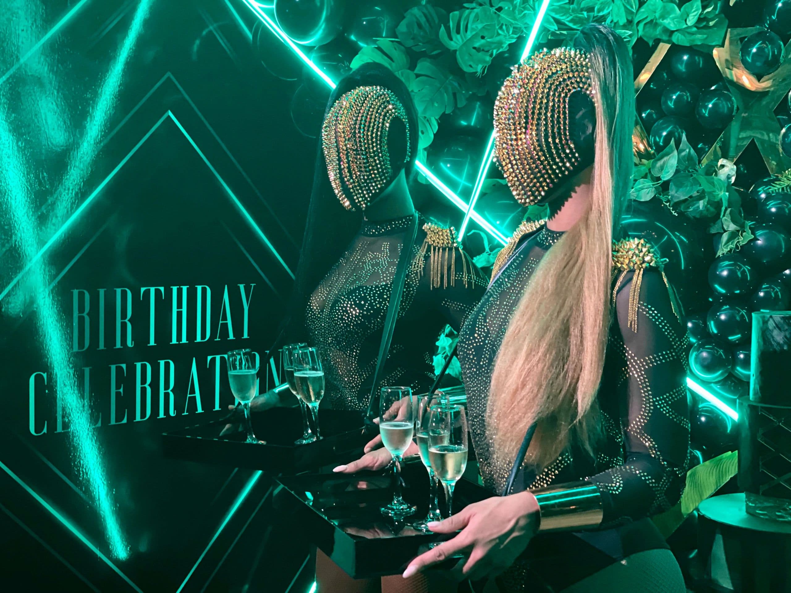 Champagne box showgirl in gold studded mask serving guests at a luxury cocktail reception
