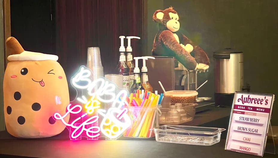 Boba tea station setup with a neon Boba Is Life sign, plush mascot, and personalized flavor menu board at a private event