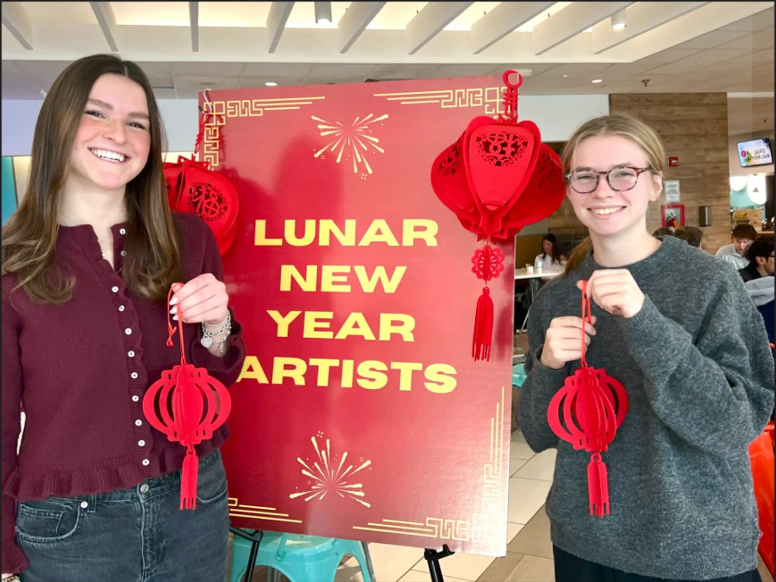 Two guests holding their finished lantern crafts in front of a Lunar New Year Artists sign at a build a lantern station