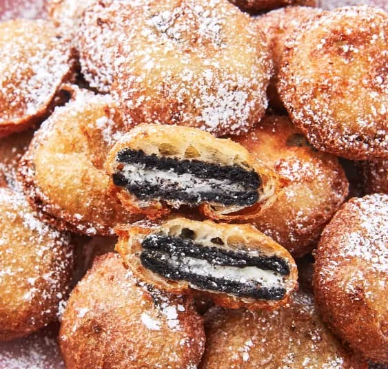 Close-up pile of deep fried Oreos dusted with powdered sugar, one split open showing the cookie center