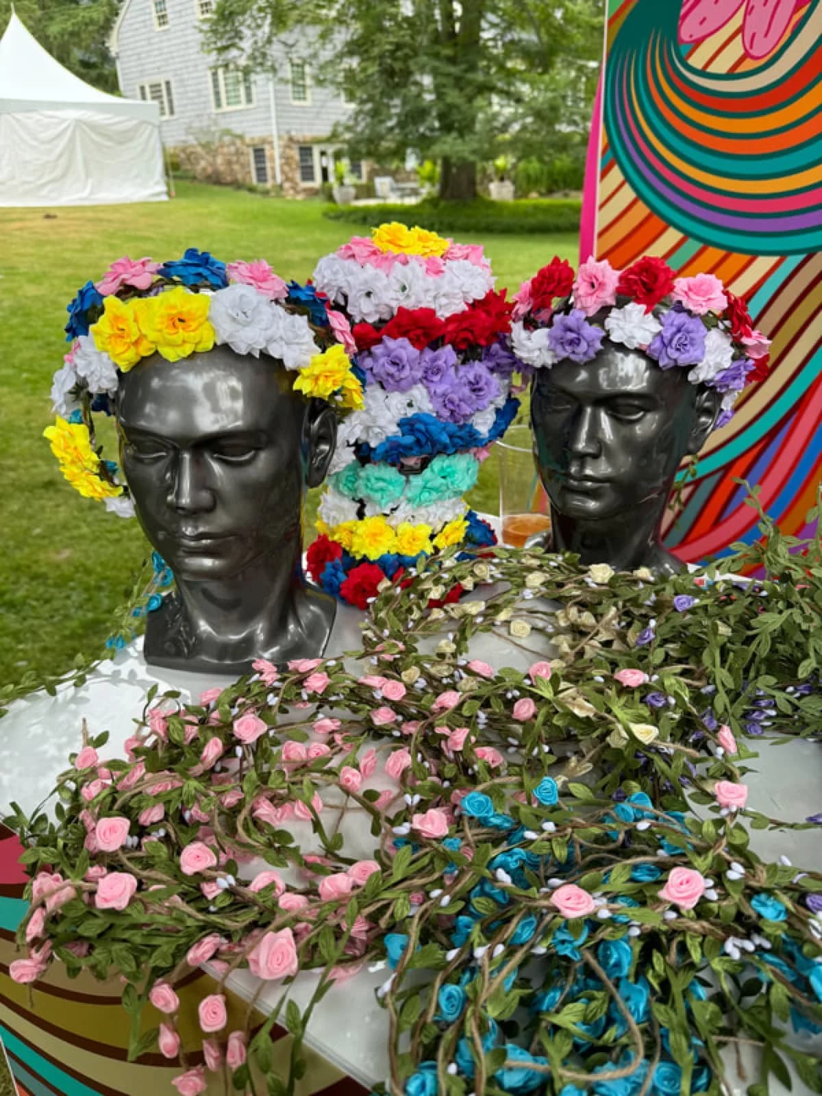 Flower crown station display with mannequin busts wearing floral crowns and bowls of silk blooms for guests to build their own at an outdoor party