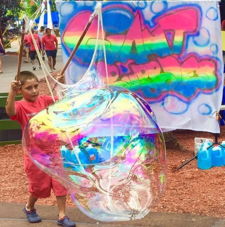 Boy holding ropes to pull a giant bubble up around himself, a giant bubble station moment at an outdoor event
