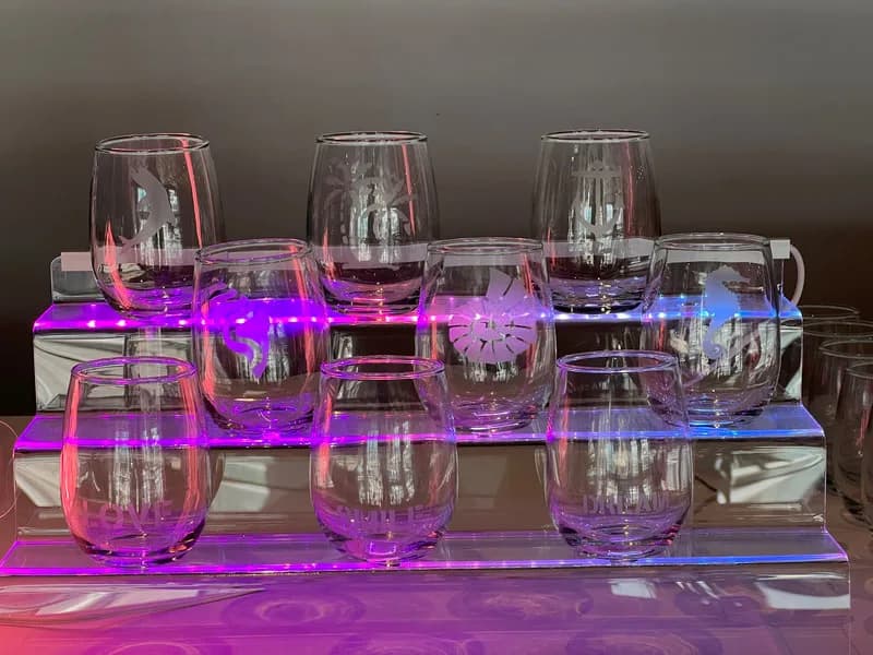 Rows of stemless wine glasses etched with dolphin, palm tree, anchor, and seahorse designs displayed on an illuminated tray at a glass etching station