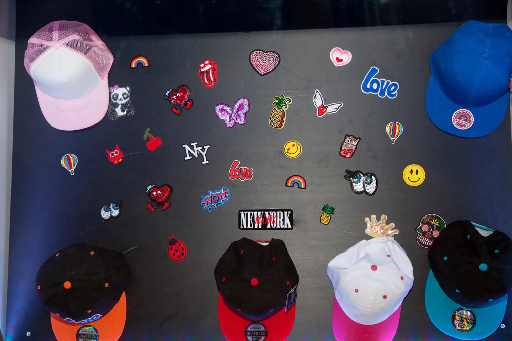 Patch and sticker display board with a colorful assortment of customizable hat decorations including hearts, emojis, and NY patches