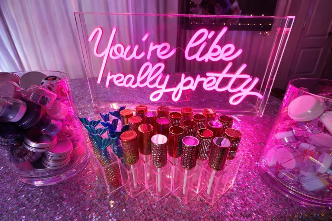 Illuminated "You're like really pretty" sign beside rows of glam lip gloss tubes at a Sweet 16 lip gloss bar