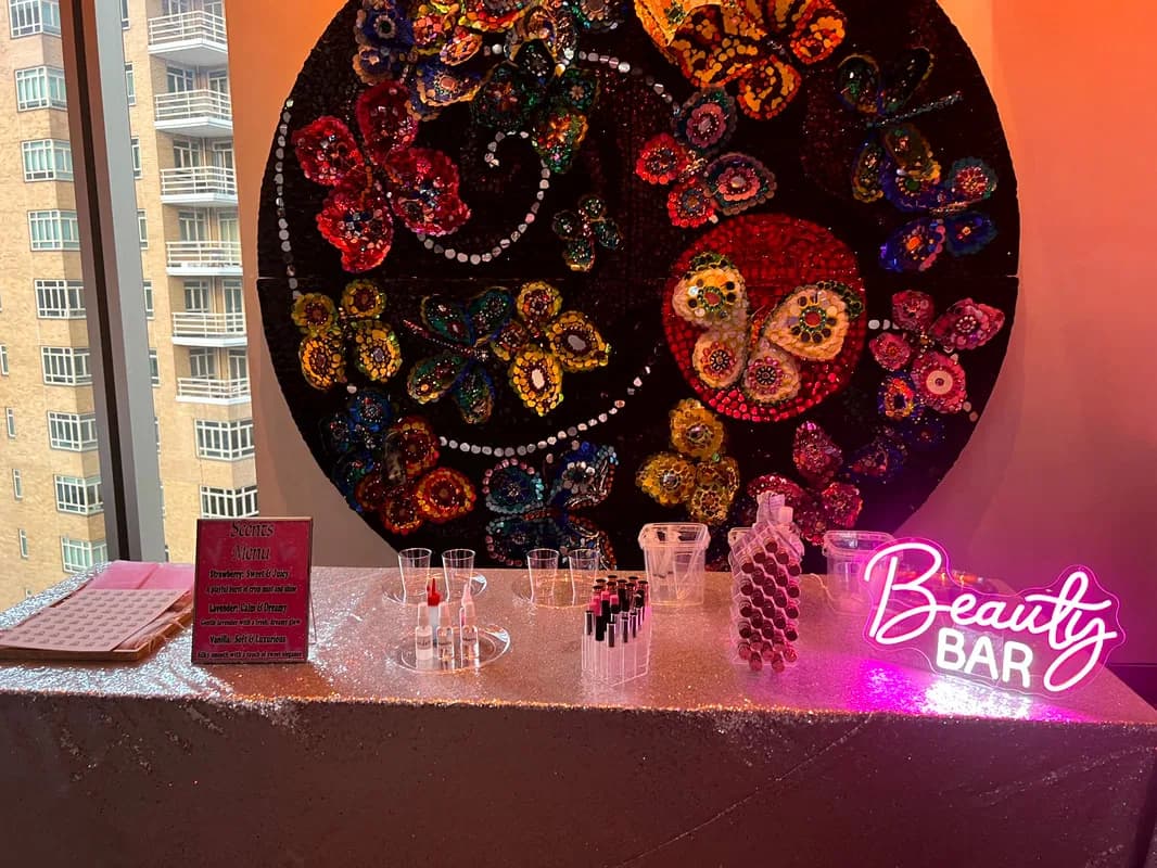 Lip gloss bar setup with a glowing Beauty Bar sign and scent menu in front of a decorative butterfly wall