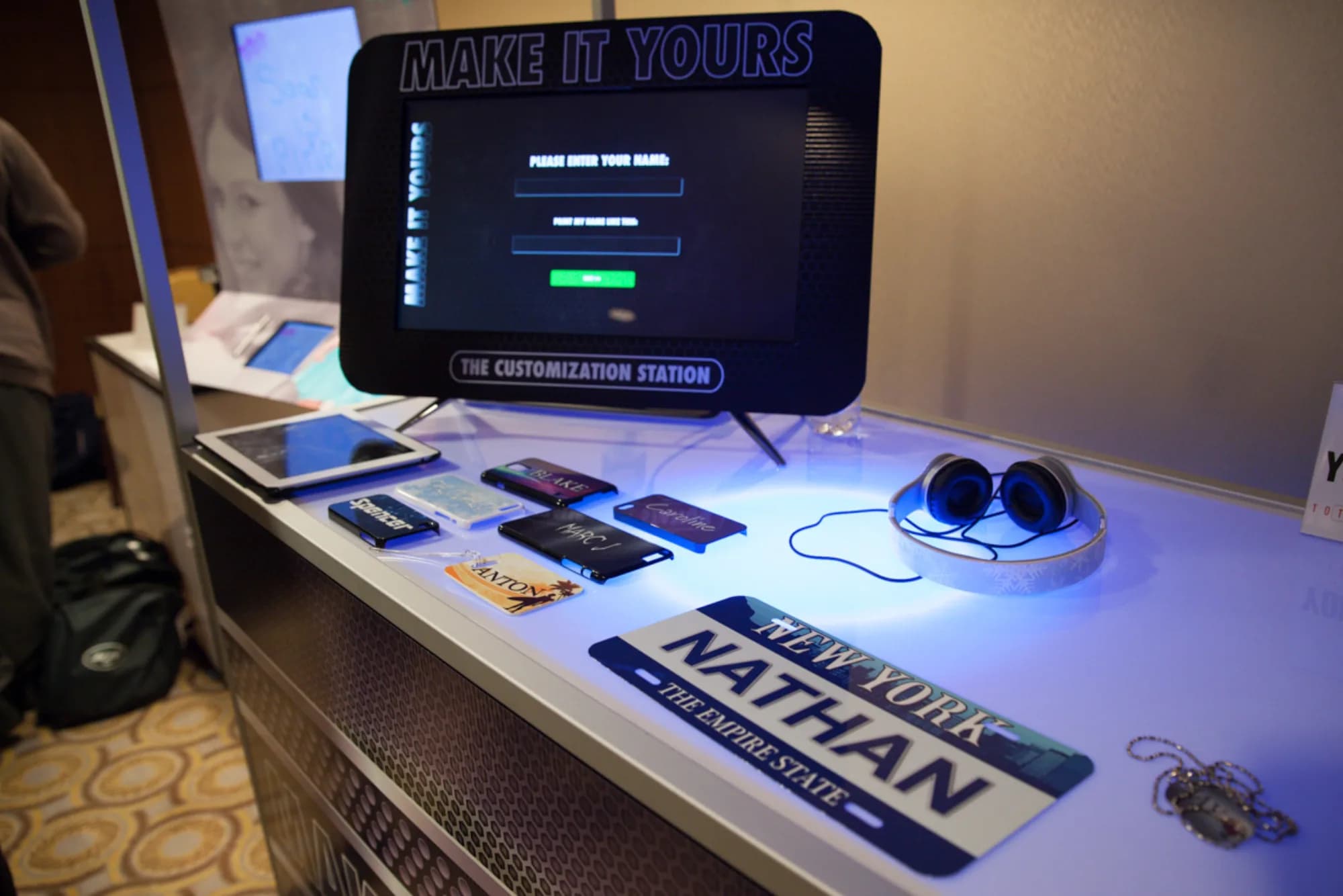 Close-up of Make It Yours kiosk table showing a spread of custom phone cases, dog tags, and a New York-themed license plate keepsake