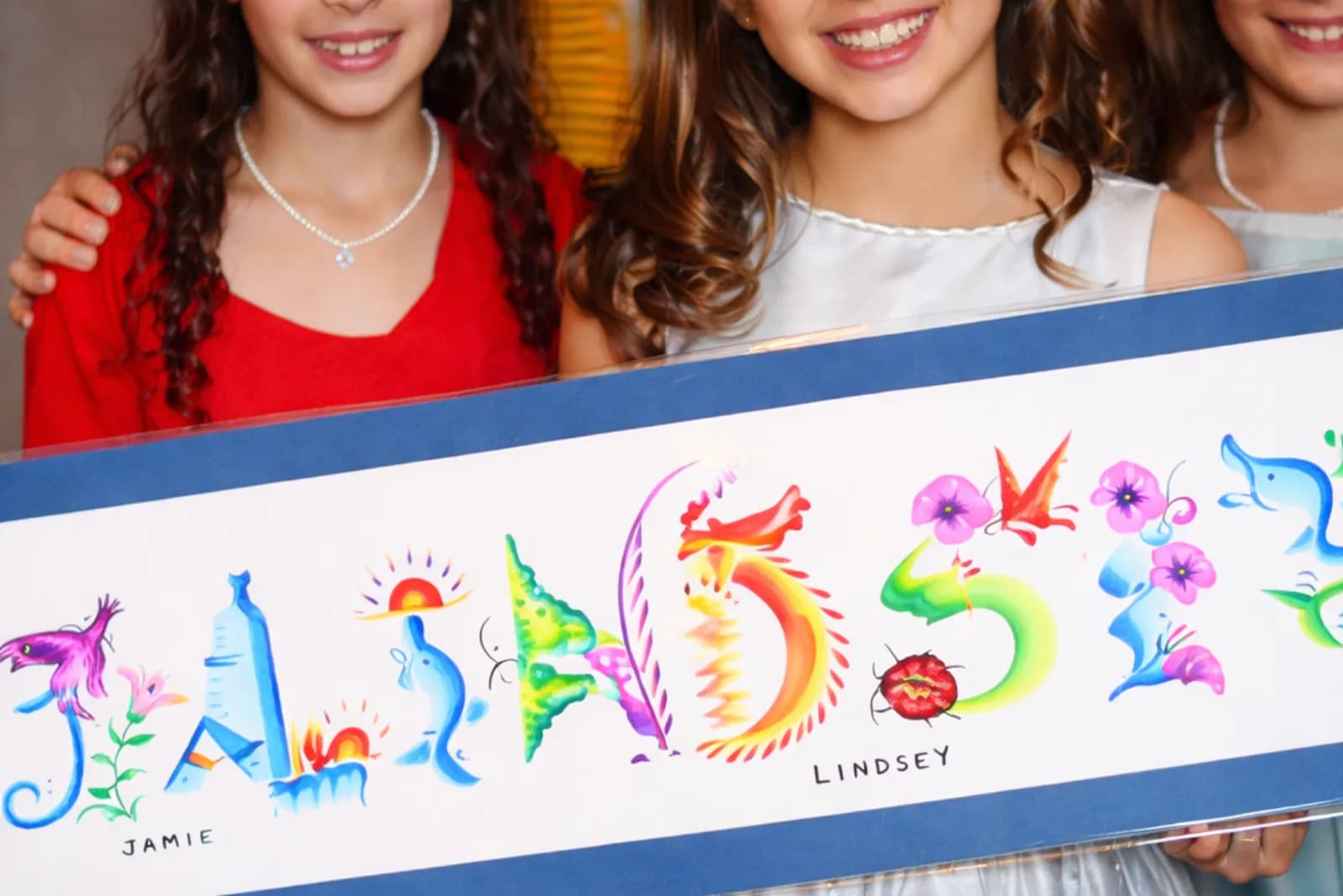Three girls holding framed name paintings spelling JAMIE and LINDSEY, each letter filled with colorful floral and animal imagery, from the name painting station