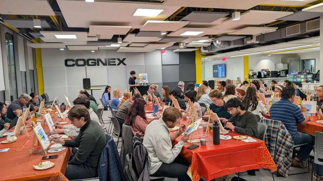 Large group of guests seated at long tables painting canvases together during an indoor corporate guided painting workshop