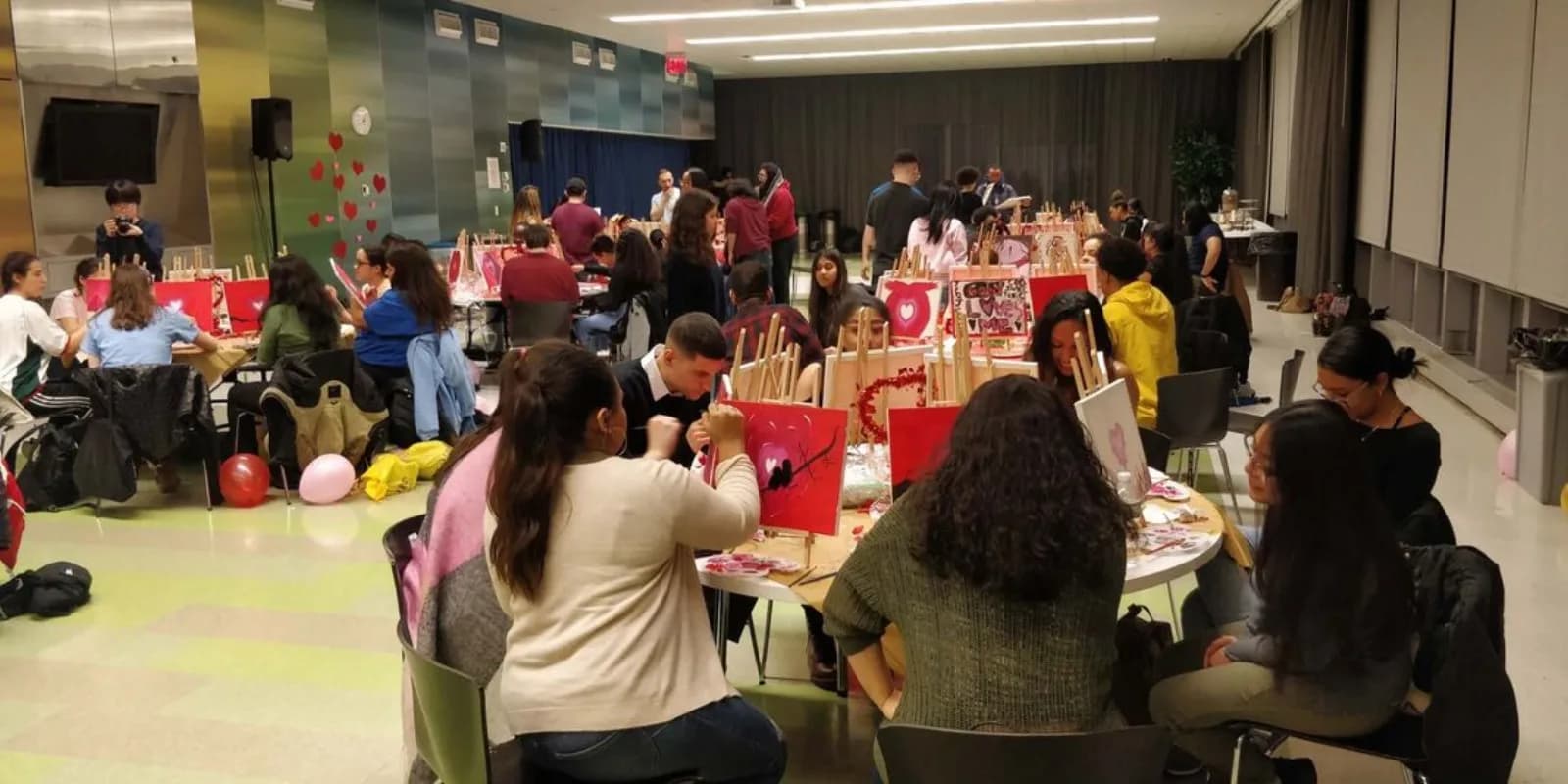 Wide view of guests seated at long tables painting heart-themed canvases together during a guided painting workshop