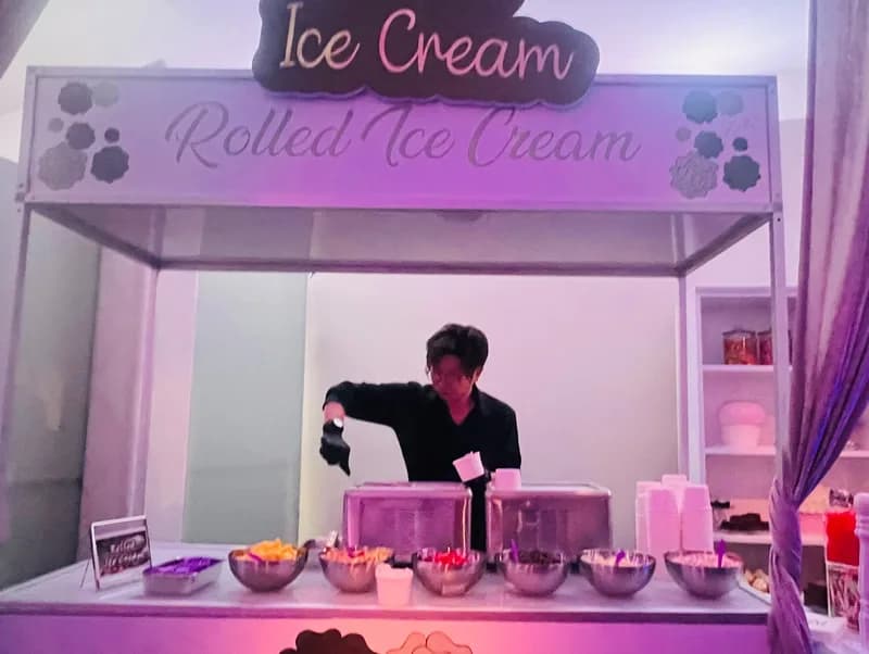 Attendant scooping toppings behind an illuminated Rolled Ice Cream kiosk sign at an evening event