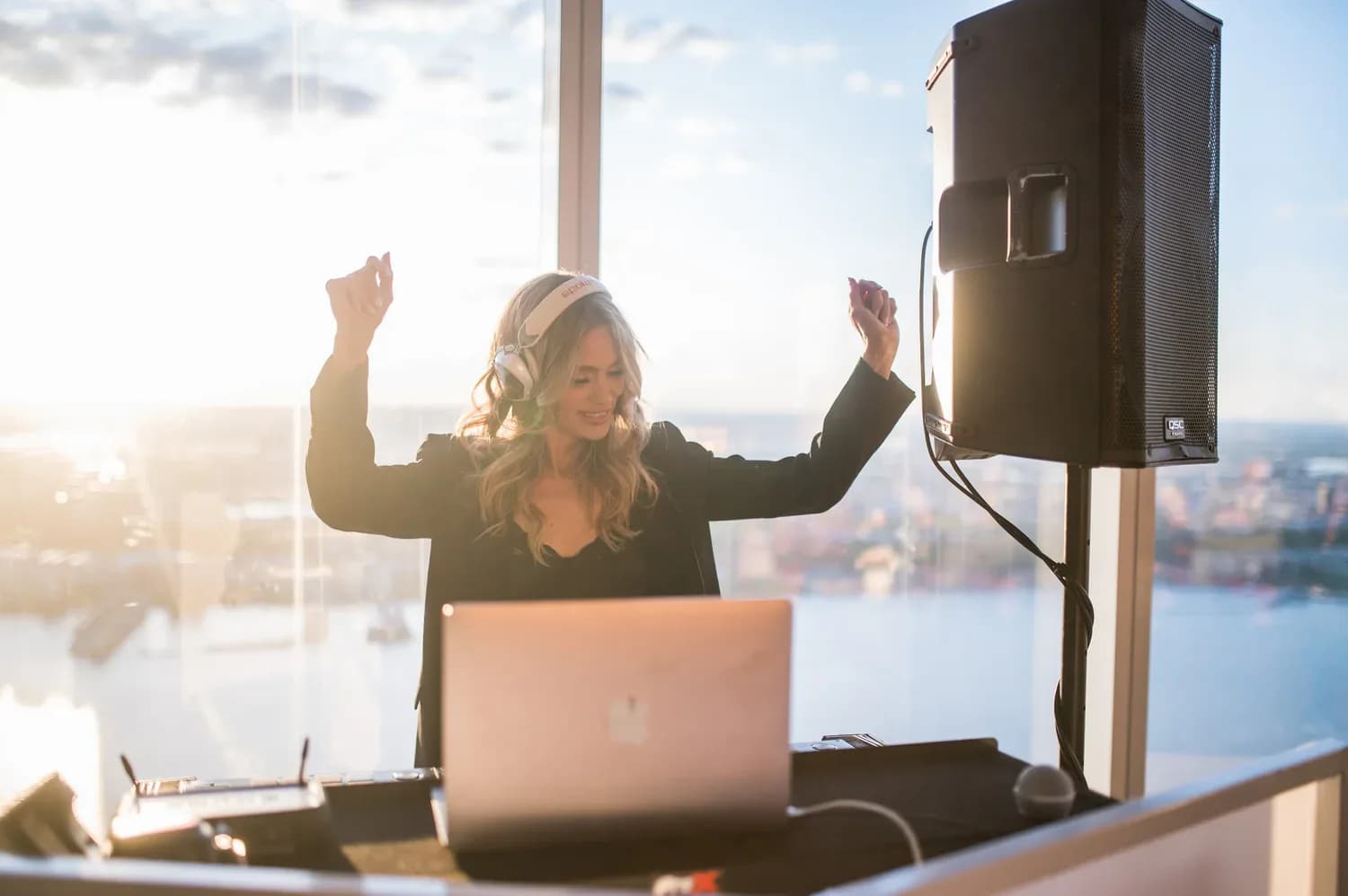 DJ Dani arms raised at the decks during a daytime high-rise event with city skyline and golden sunlight