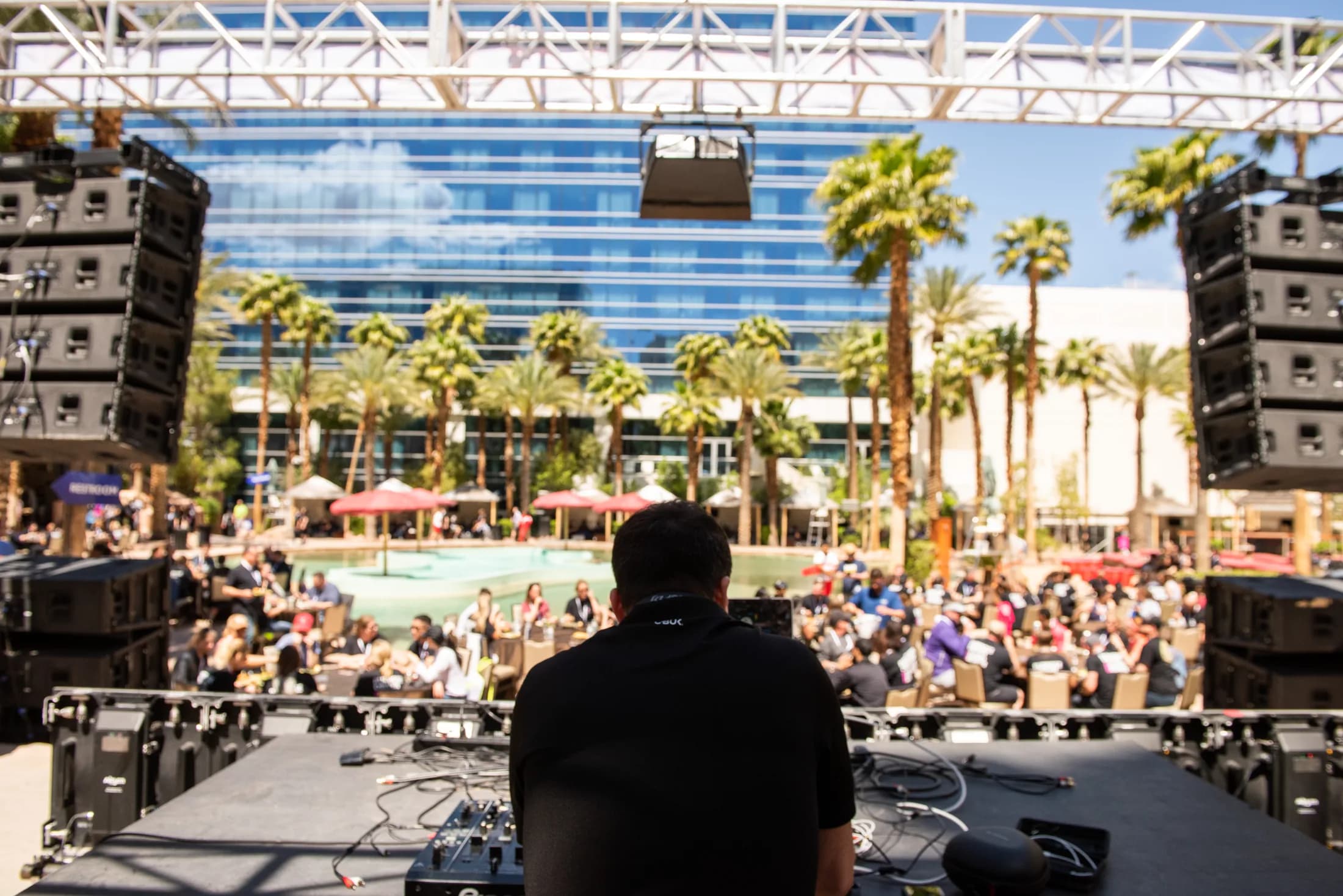 A large open-air rooftop festival stage surrounded by palm trees and hundreds of guests