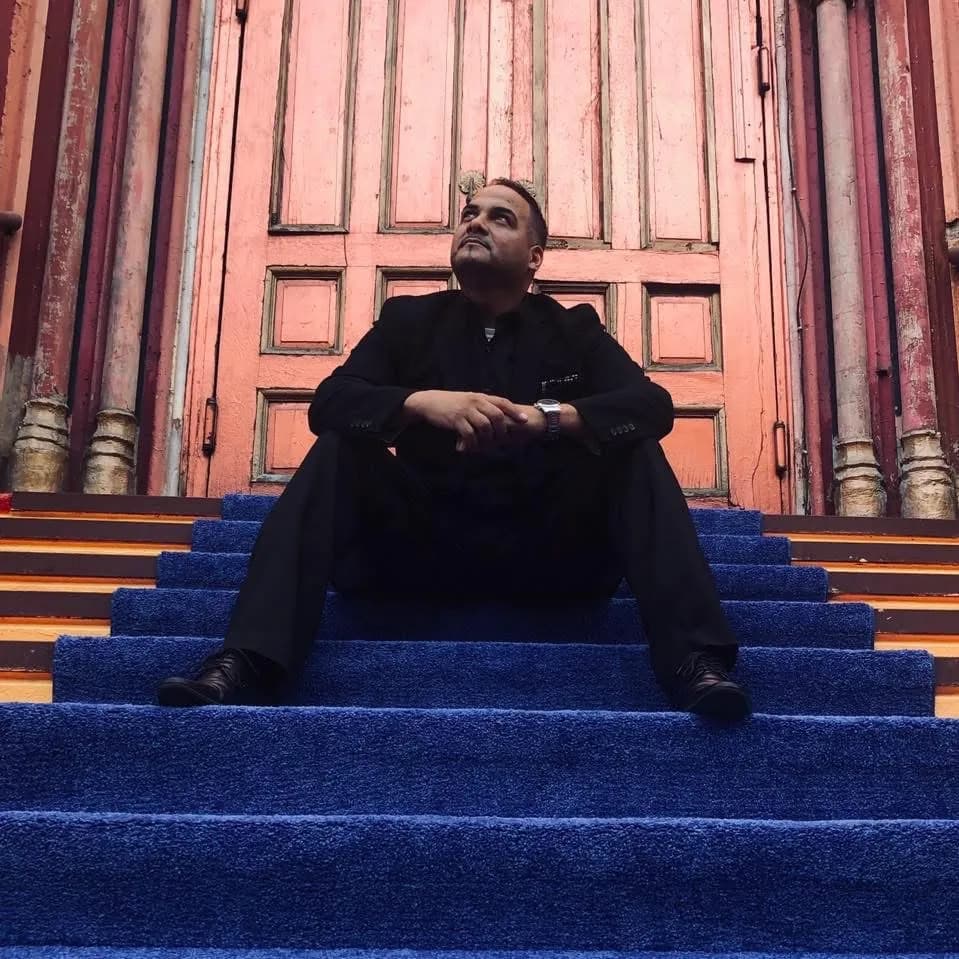 Omar in a black suit seated on blue carpeted steps in front of tall weathered doors, editorial portrait