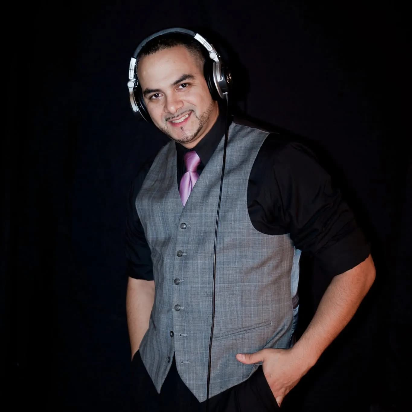 Omar in a grey waistcoat and tie wearing headphones, studio press portrait on a black backdrop