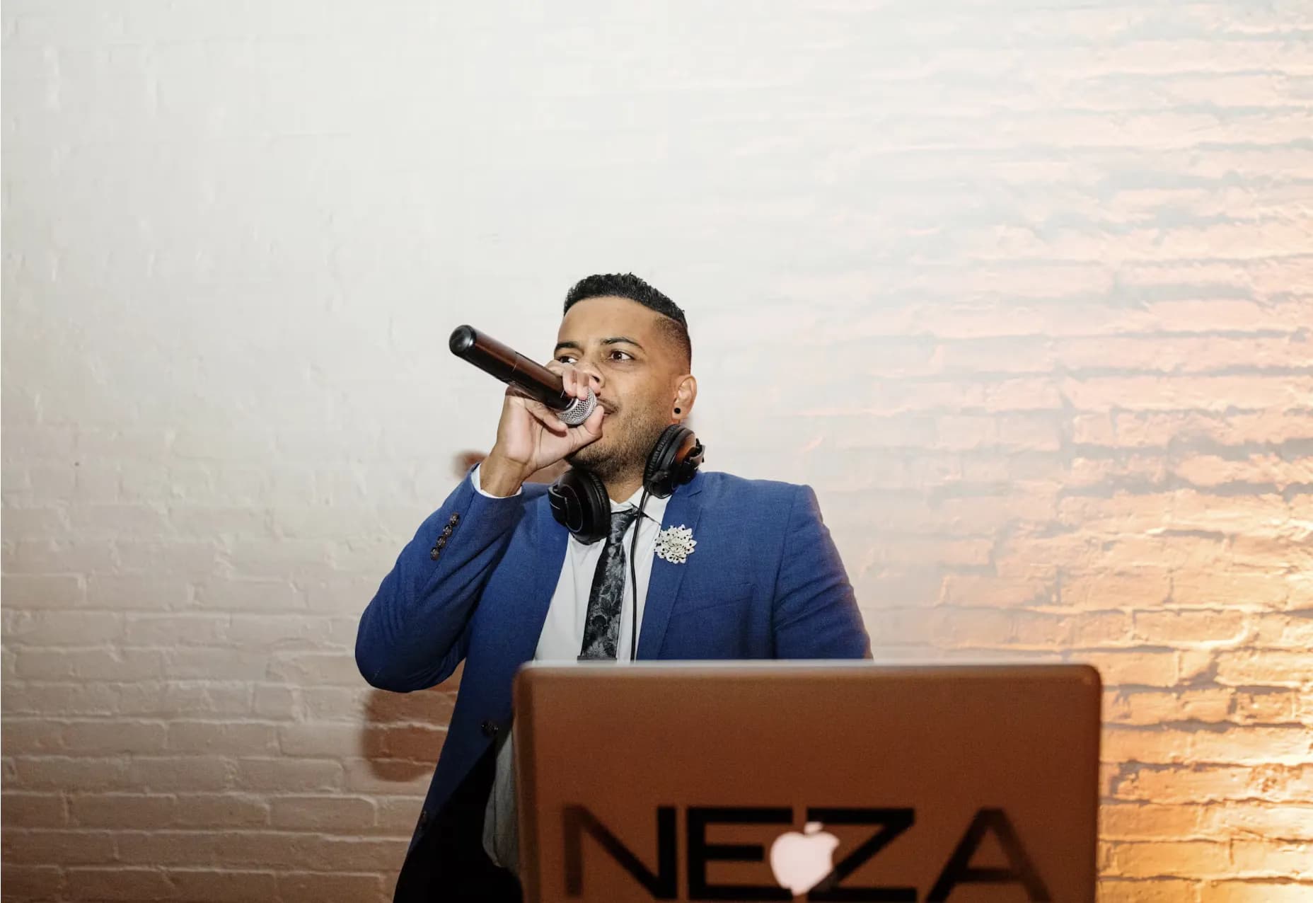 DJ Neza in a blue jacket on the microphone hosting at an event with warm uplighting