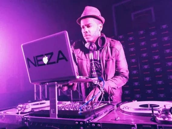 DJ Neza behind the turntables in a hat, his NEZA laptop open, purple stage lighting illuminating the set