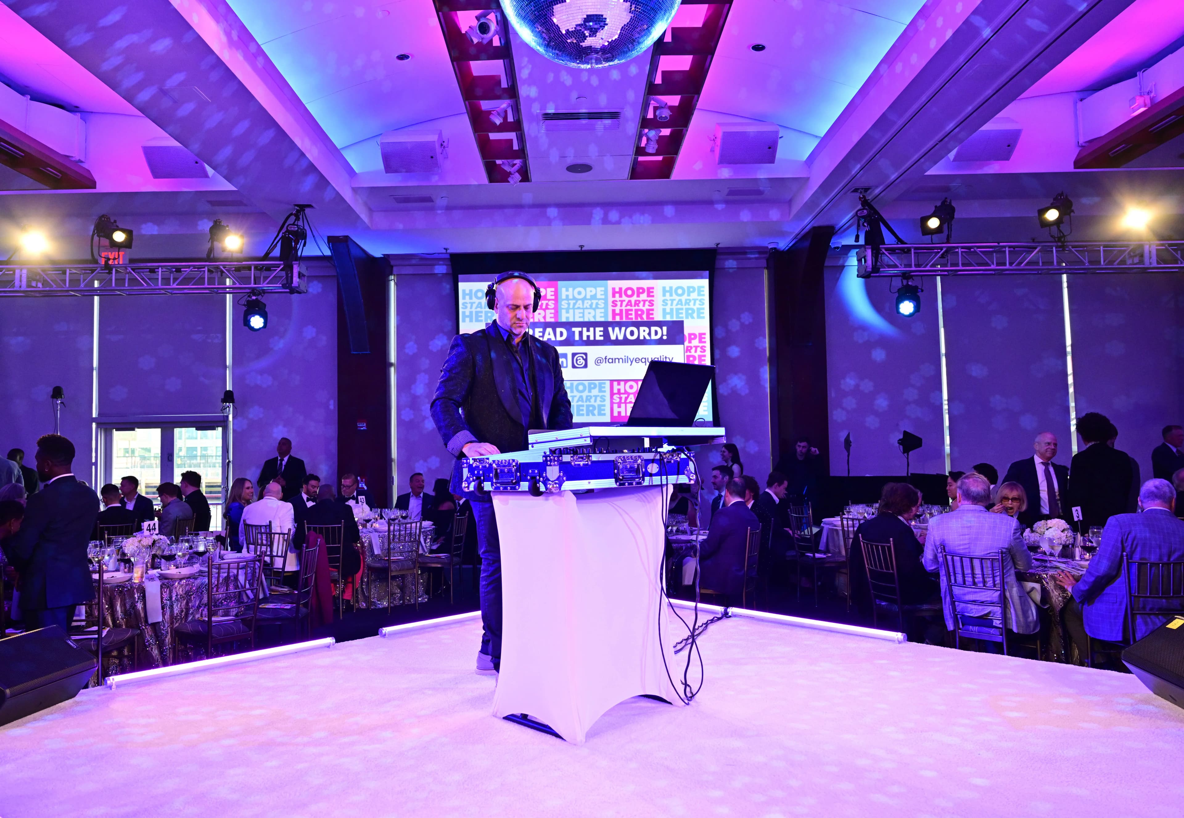 Shiran Nicholson at the DJ booth with a packed white dance floor and production screens at a gala fundraiser event