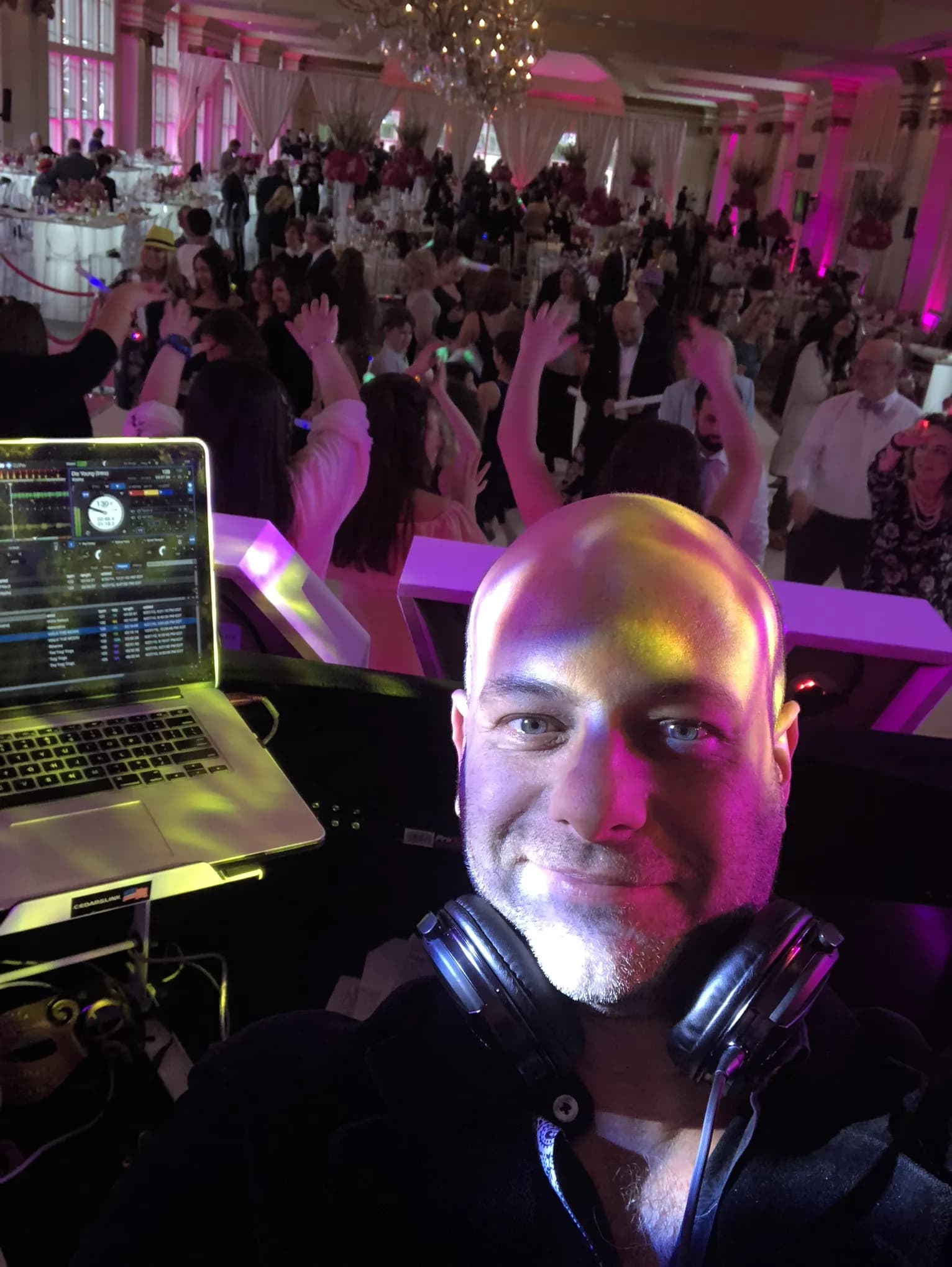 DJ Shiran Nicholson at the booth with a packed pink-lit ballroom and dancing crowd behind him at a celebration