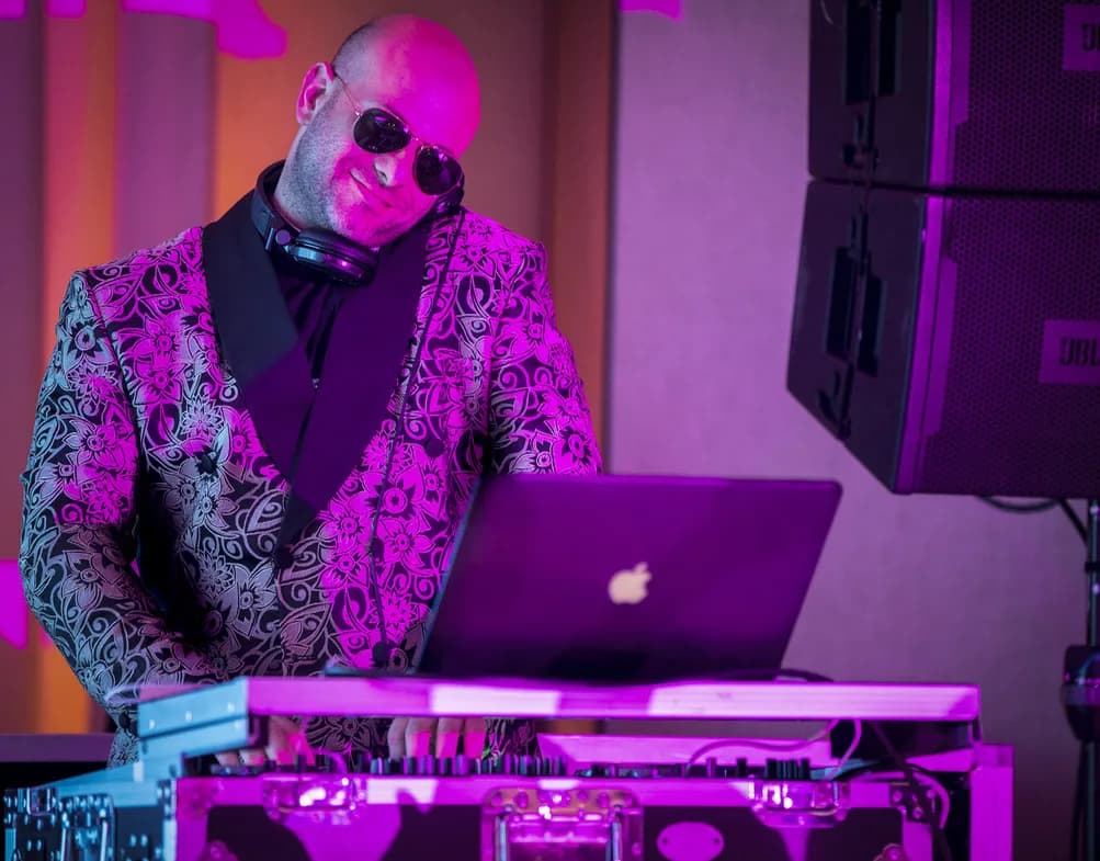 DJ Shiran Nicholson in a pink paisley jacket and sunglasses at the decks under magenta lighting at an event