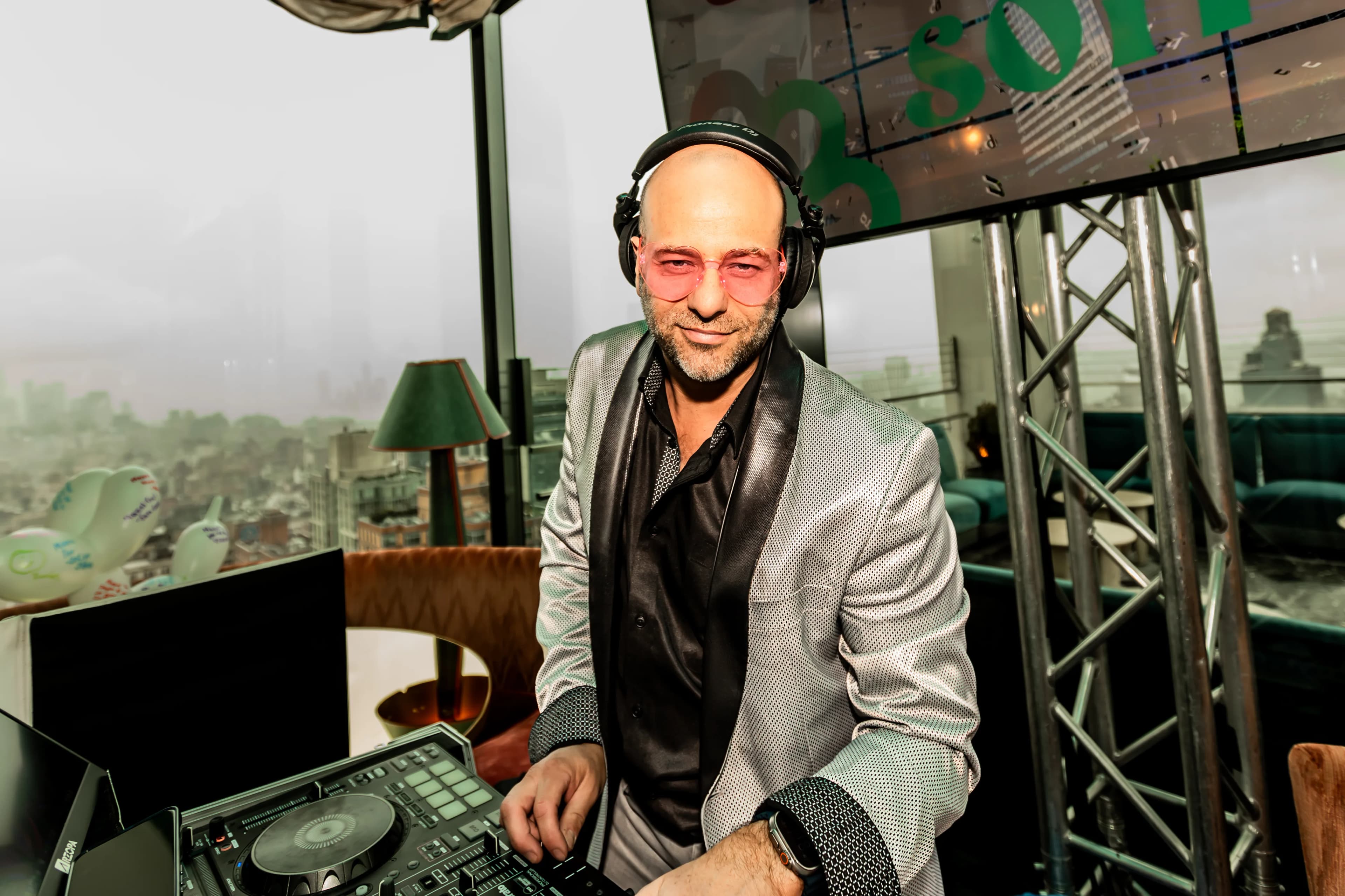 DJ Shiran Nicholson in a silver blazer and headphones at the decks on a rooftop venue overlooking the city