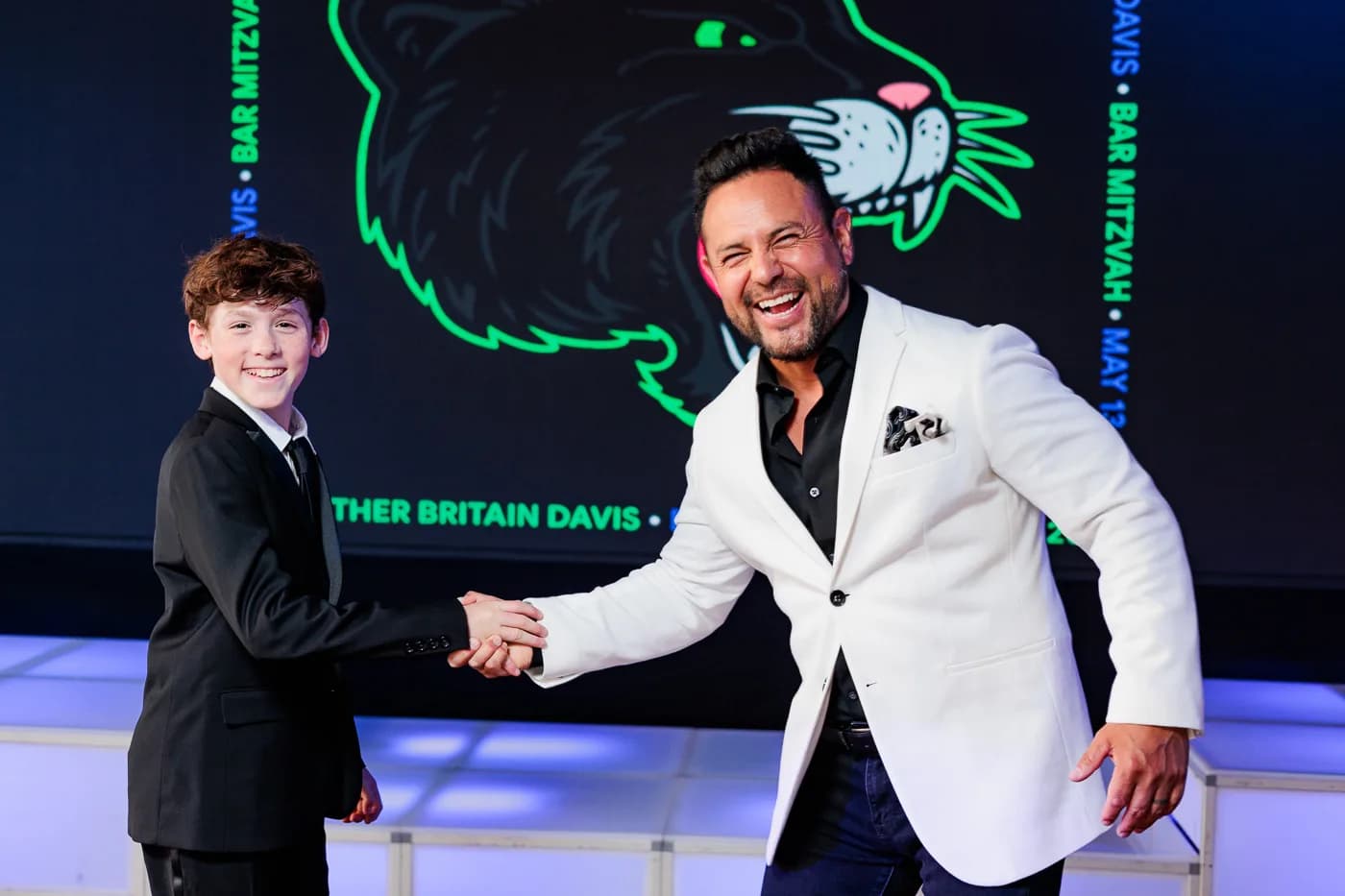 MC Nelson in a white jacket sharing a beaming handshake with the bar mitzvah boy on a neon-lit stage