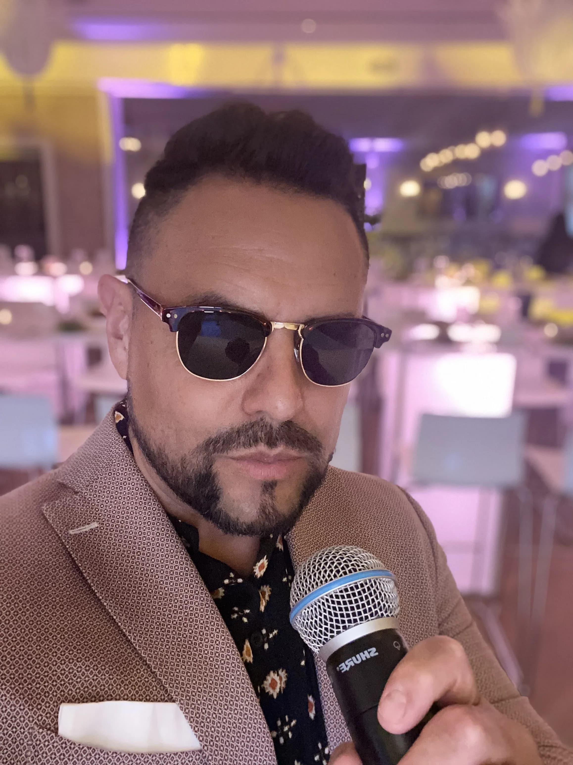 MC Nelson close-up with Shure microphone at a warmly lit event venue, sunglasses, poised to host
