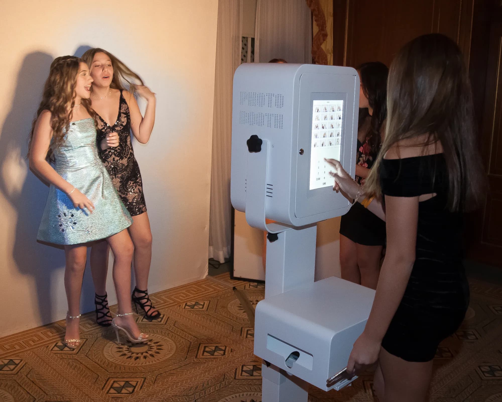 Teenagers using a white open-touchscreen photo kiosk at a formal event with a printed strip in the output tray