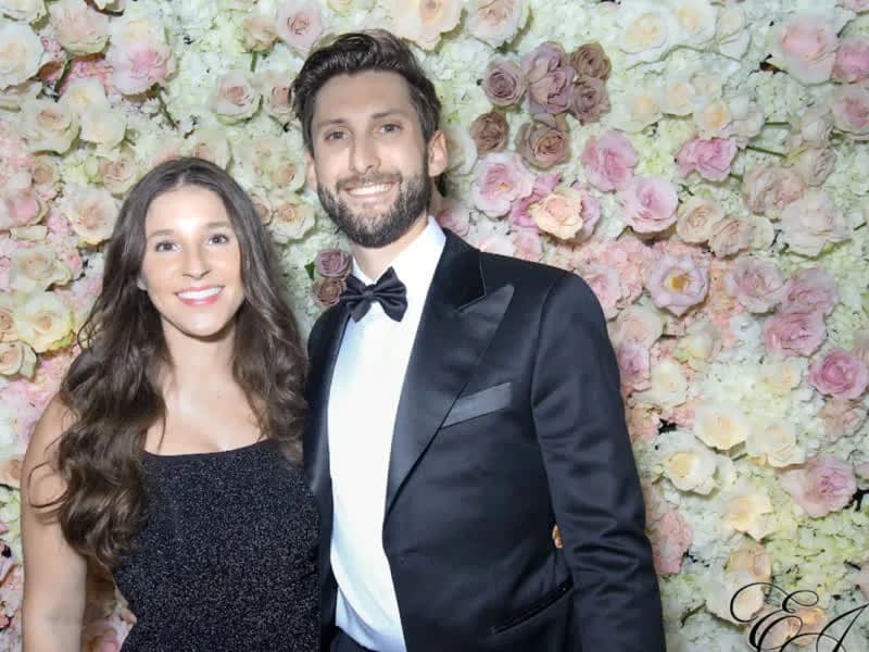 Couple in formal attire posing in front of a flower wall backdrop at an evening celebration