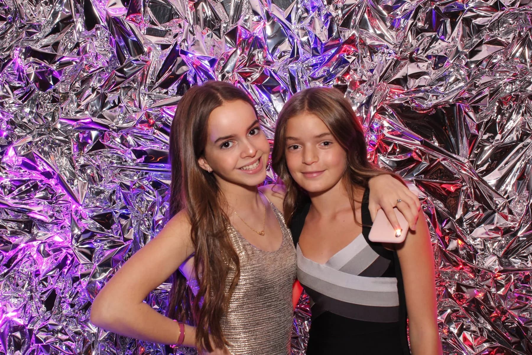 Two guests posing in front of a silver crinkle foil backdrop with pink and purple uplighting at a New York event