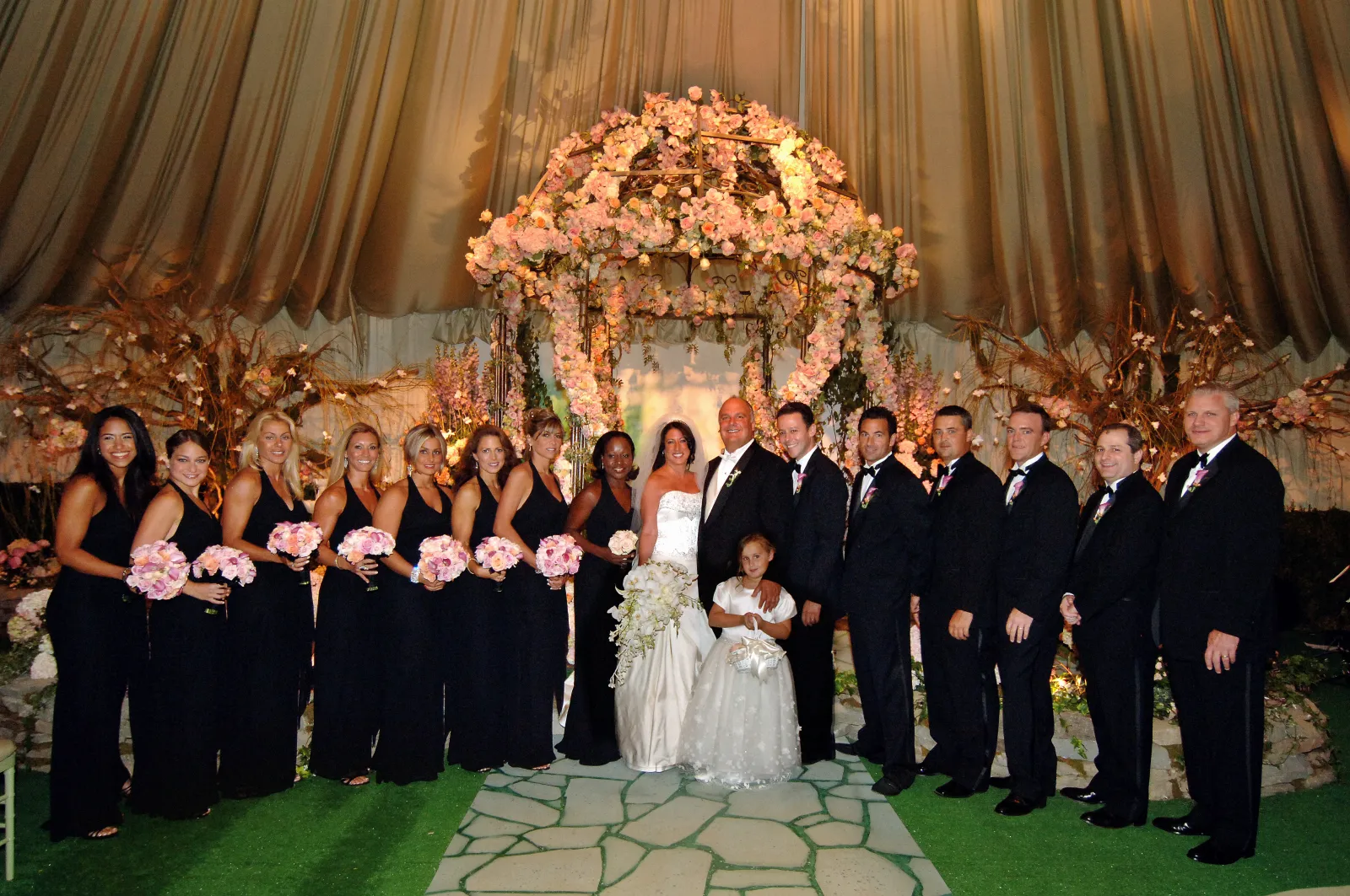 Bride, groom, and wedding party posed beneath a flower-covered arch inside a warmly lit tent at a wedding produced by Nicholson Events